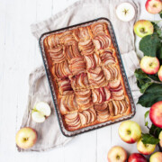 Square shaped apple tart in glassware pan. A flatlay image on white wooden background. Fresh apples, apple leaves and white linen.