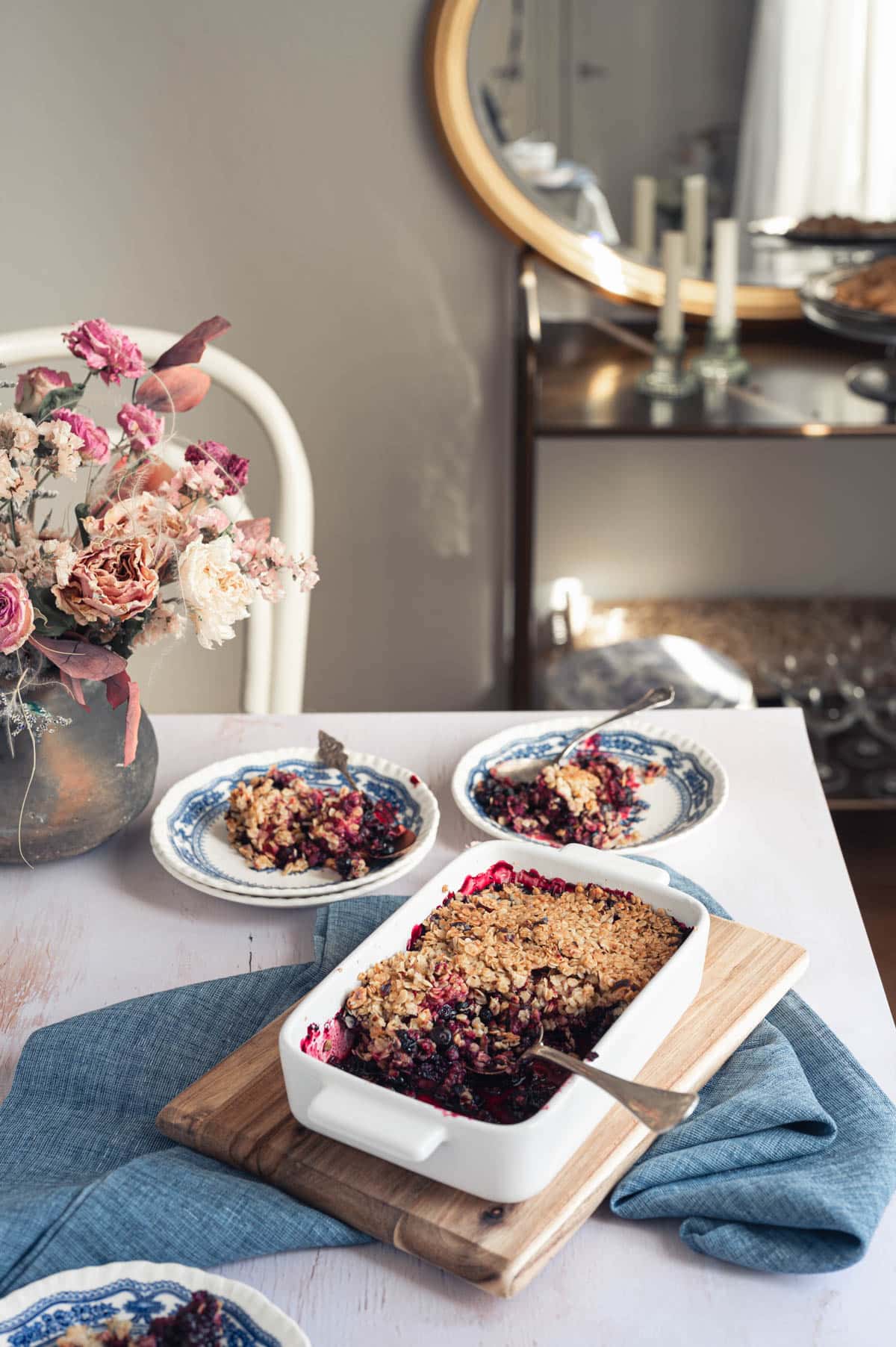 Berry crisp served on the table. The crisp is in a white baking dish. It has been portioned onto blue-patterned plates. There are dried flowers in a vase on the table, and in the background, you can see a serving trolley and a mirror.
