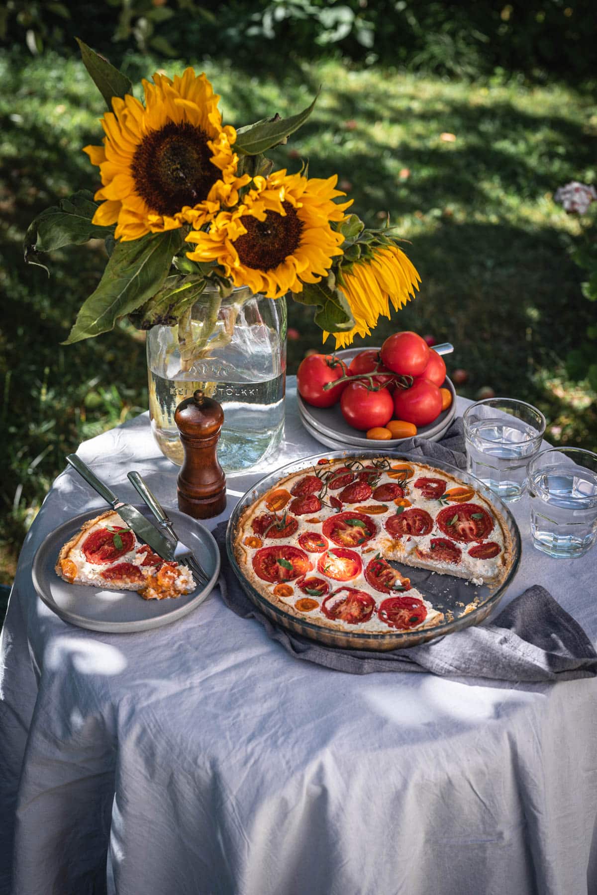 Tomaattipiirakka puutarhapöydällä. Piirakka on pyöreässä piirakkavuoassa ja siitä on leikattu yksi pala harmaalle lautaselle. Pöydällä on myös auringonkukkia maljakossa sekä tuoreita tomaatteja lautasella.